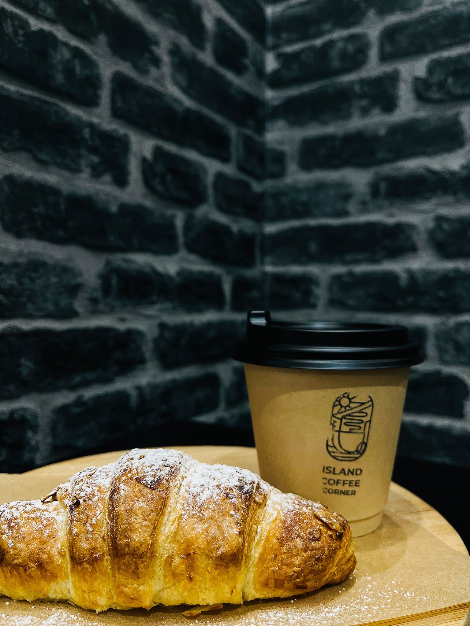 Croissant dusted with icing sugar beside a branded Island Coffee Corner takeaway cup