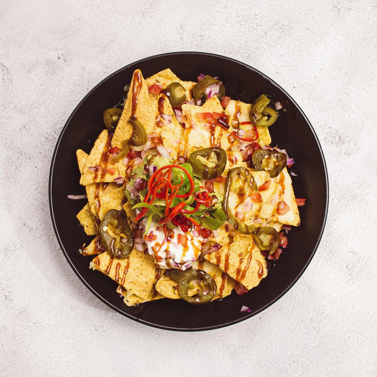 Nachos in black bowl with sour cream, jalapenos, pico de gallo and chilli drizzle