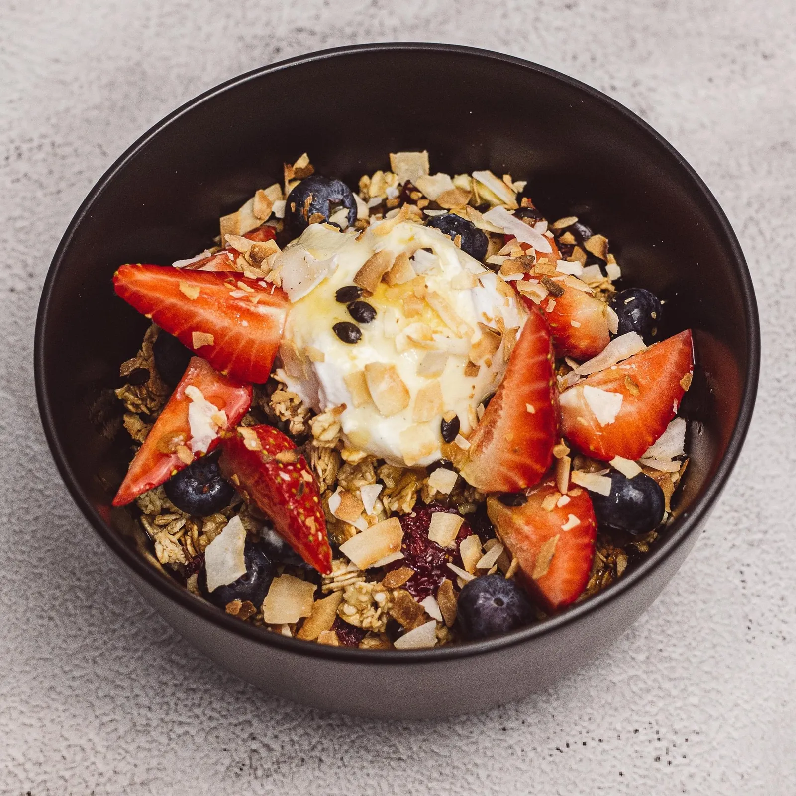 Granola bowl with strawberries, blueberries, coconut and honey yoghurt in black ceramic bowl